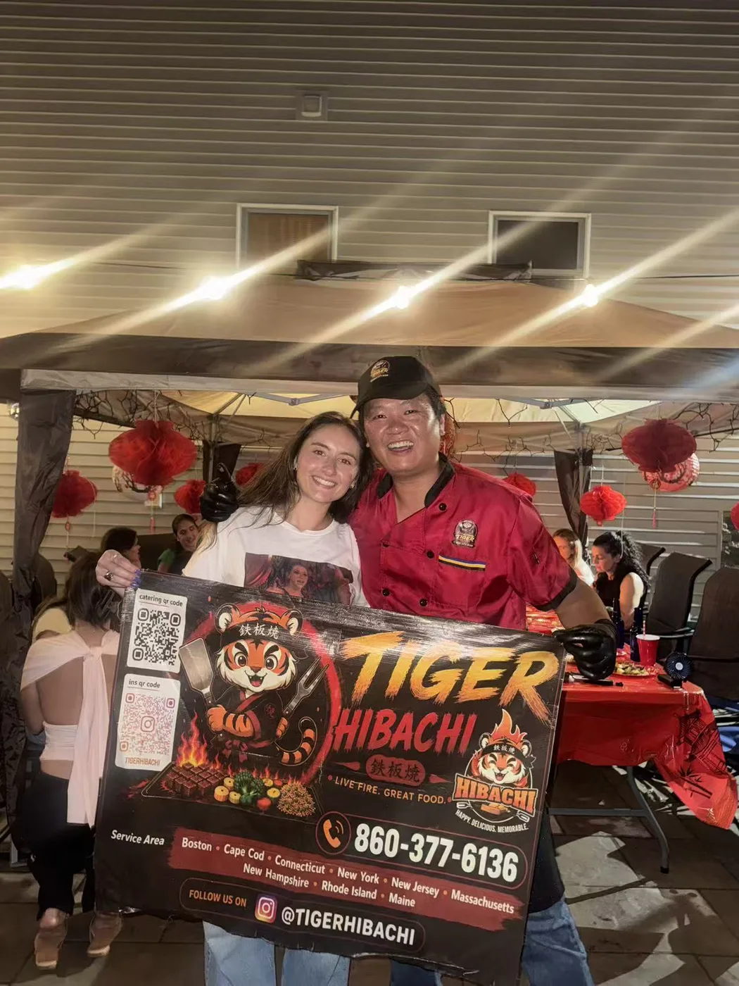 Tiger Hibachi chef in a red chef coat posing with a guest and the Tiger Hibachi banner at an evening Connecticut party